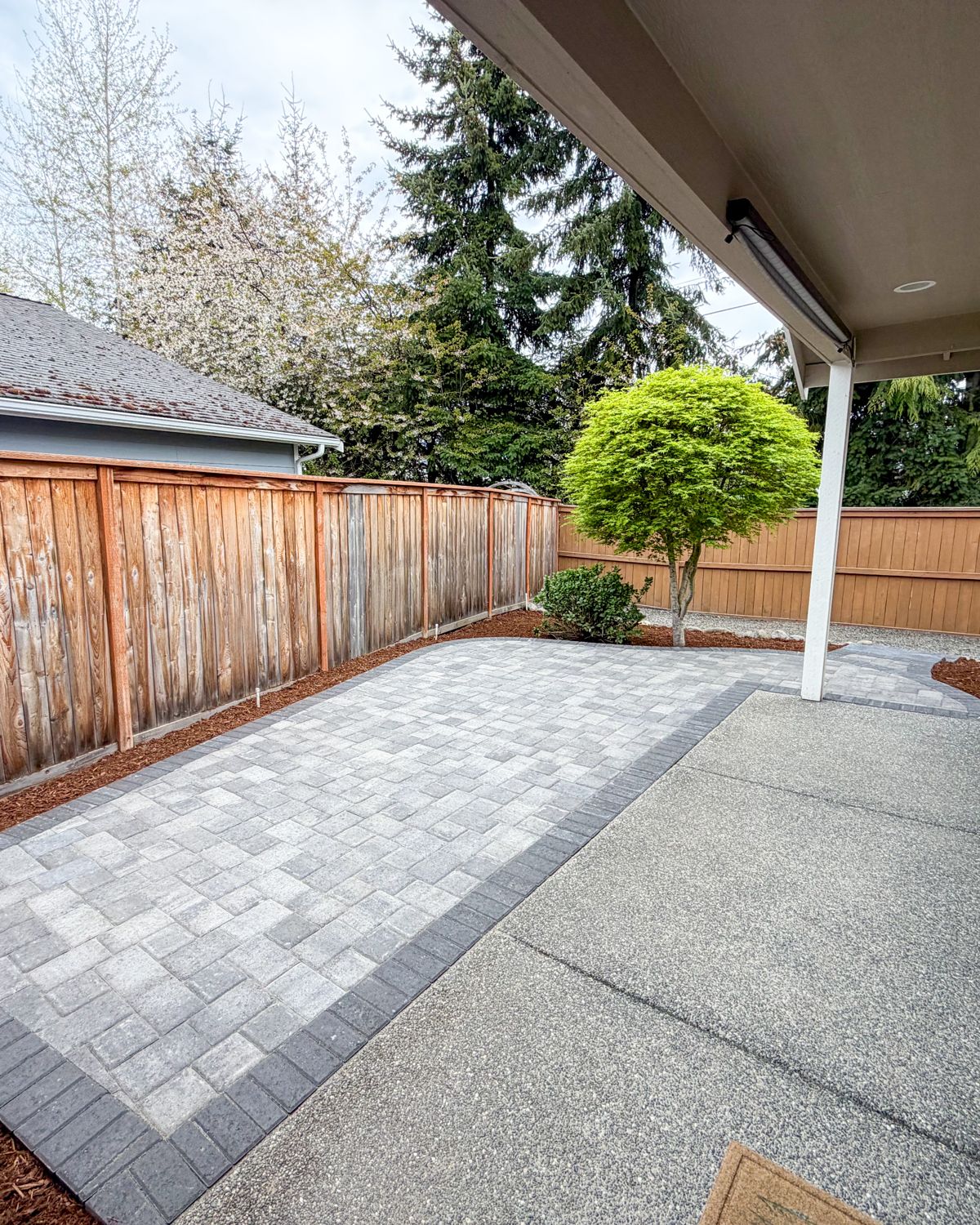 Gray paver patio with a charcoal border off a covered porch next to a Japanese maple