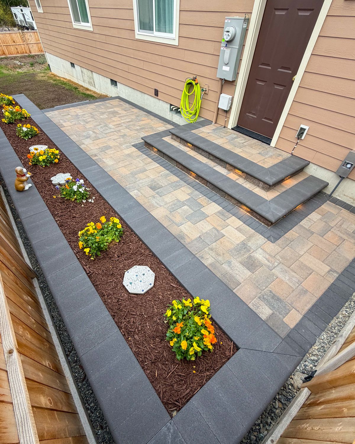 Paver patio with lit steps and a bordered flower bed beside the back door